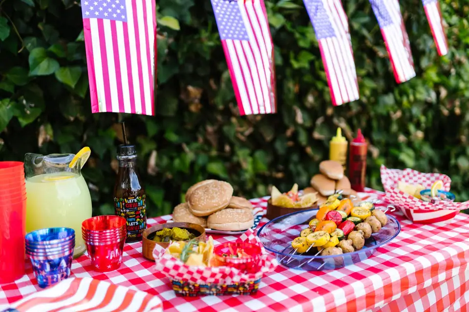 A picnic table with a red checked cloth, burgers, drinks and small American flags under flag bunting