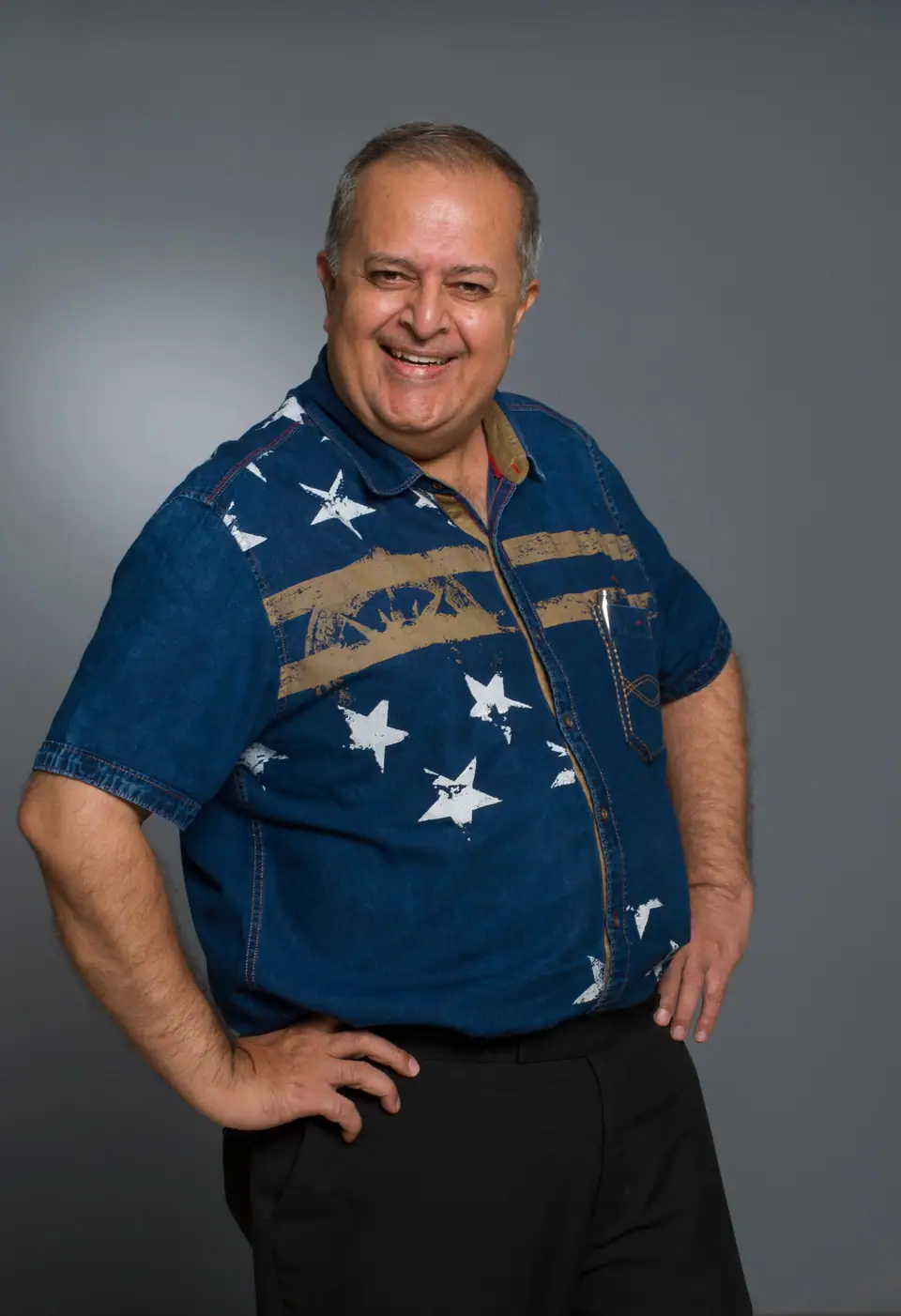 A smiling man in a blue denim shirt printed with white stars, hands on his hips