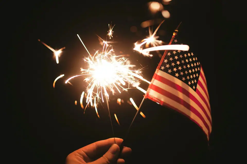 A hand holding a lit sparkler and a small American flag at night